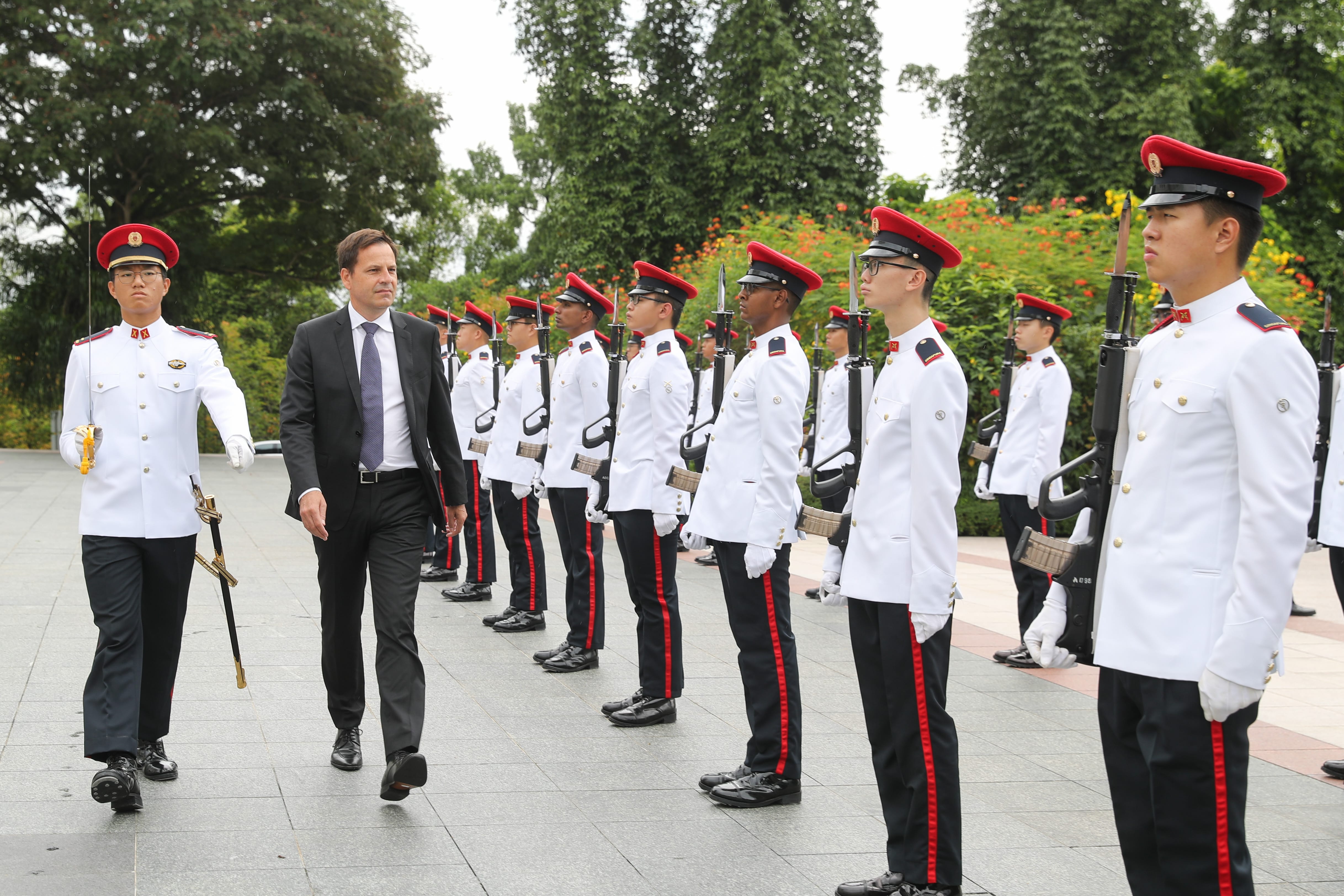 Man in suit walks past a military honor guard with rifles in white uniforms.